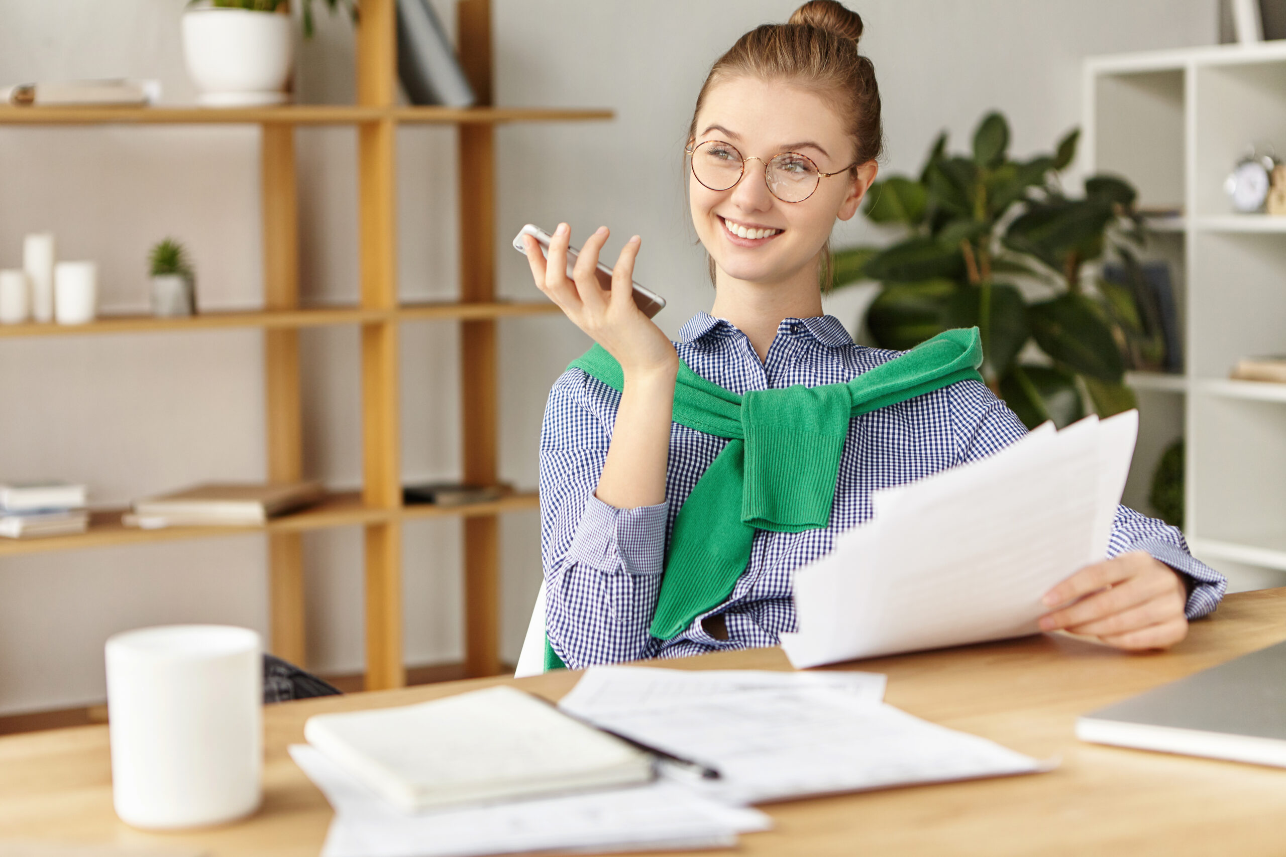 Про нас Young talented female office worker sits in cozy cabinet, holds smart phone as going to make call, keeps papers, works on expense report, has happy expression, notices something pleasant outside