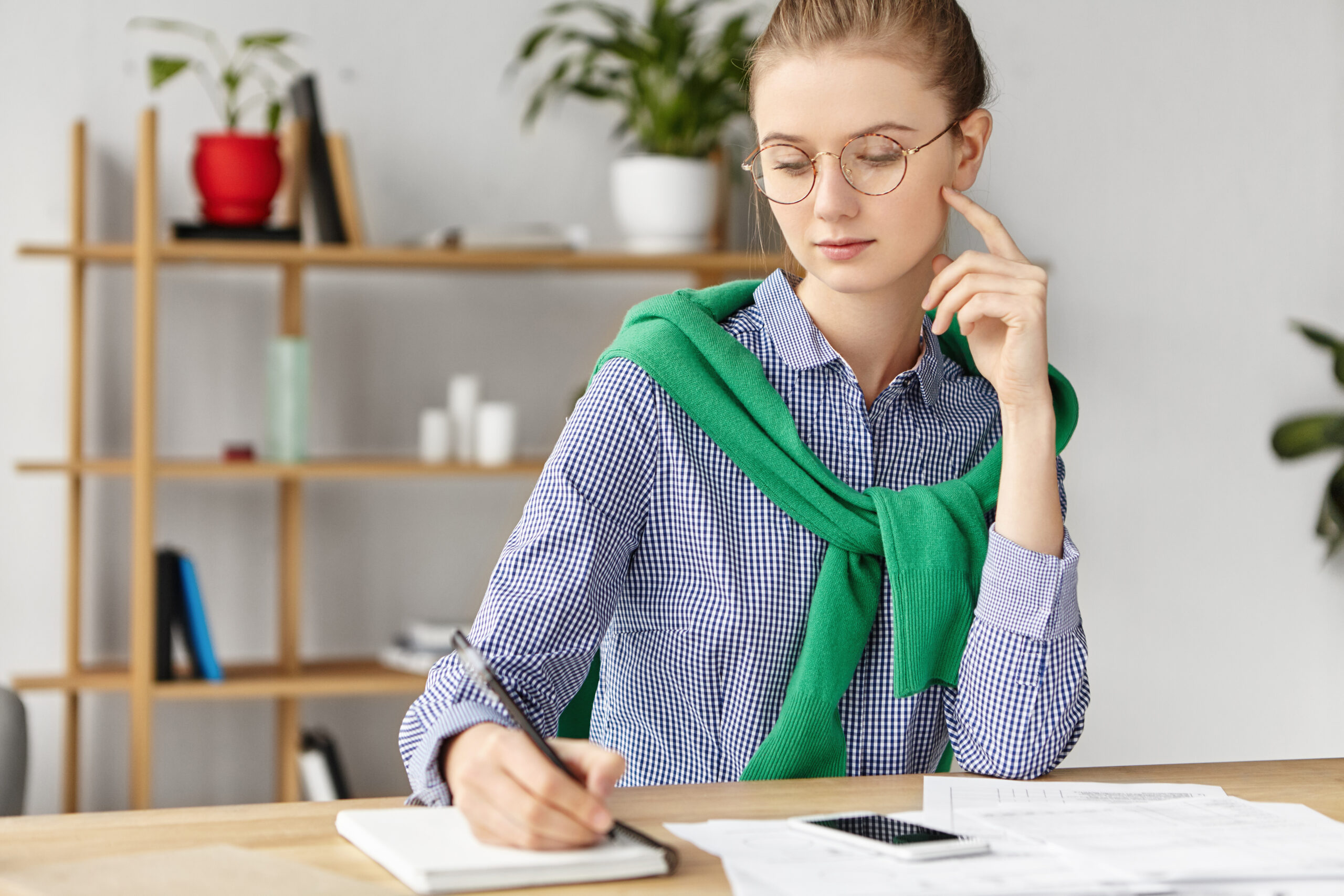 Наші роботи Indoor shot of attractive young female wears checkered shirt, writes at notebook, makes lists of meetings and arrangements for whole working weekend, sits in cozy office, focused on writing.